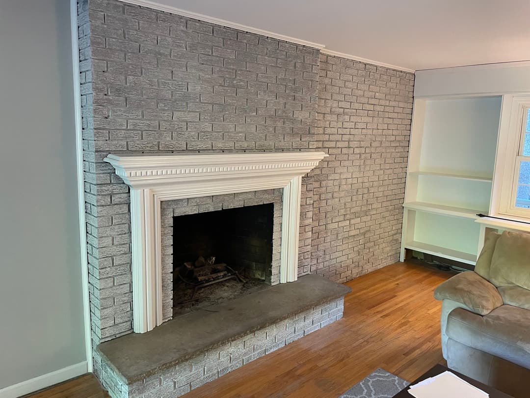 Fireplace with white mantel on brick wall in a cozy living room with wooden floor and beige couch.