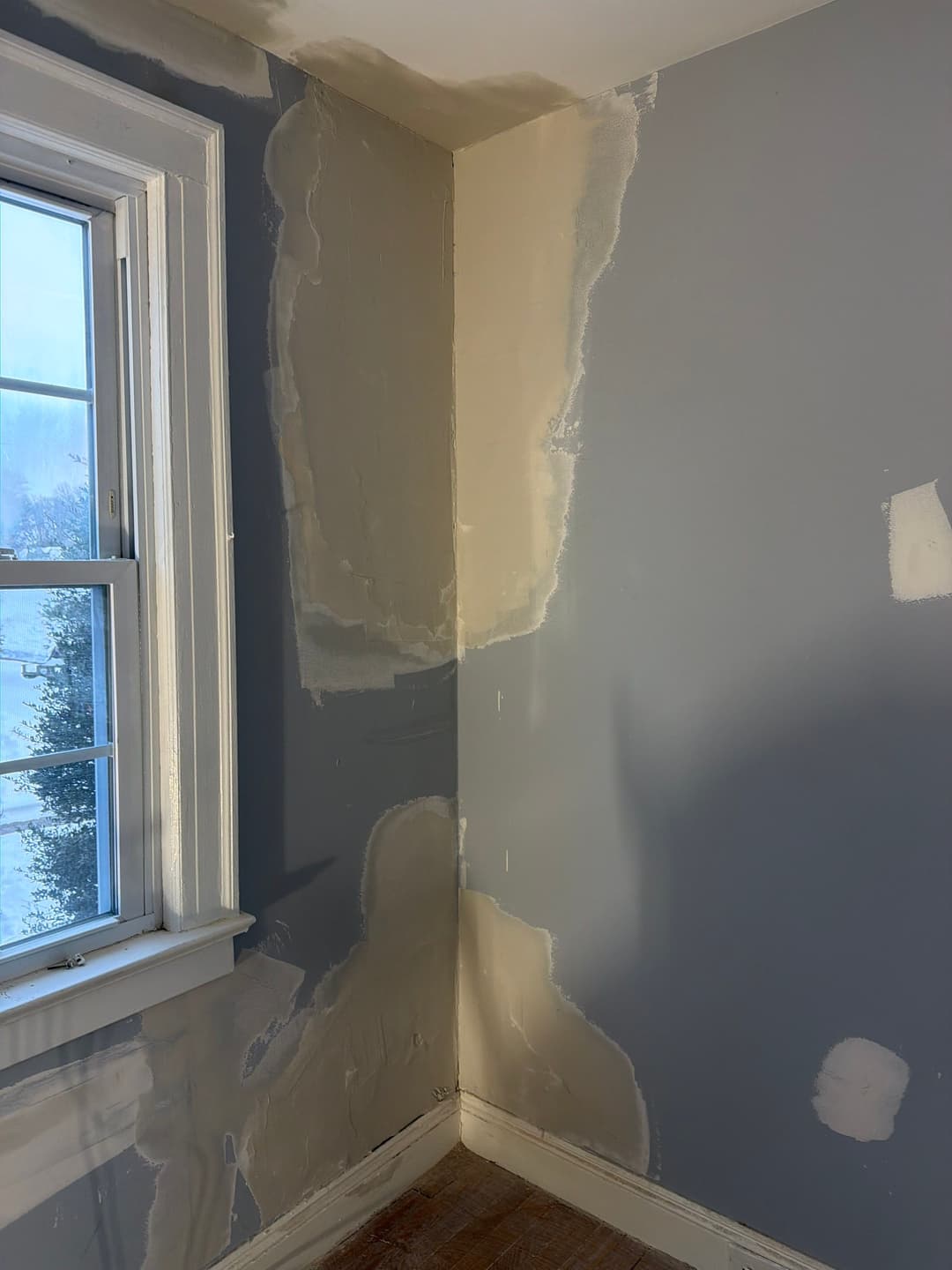 Interior corner of a room with patched gray walls and a window, showing renovation progress.