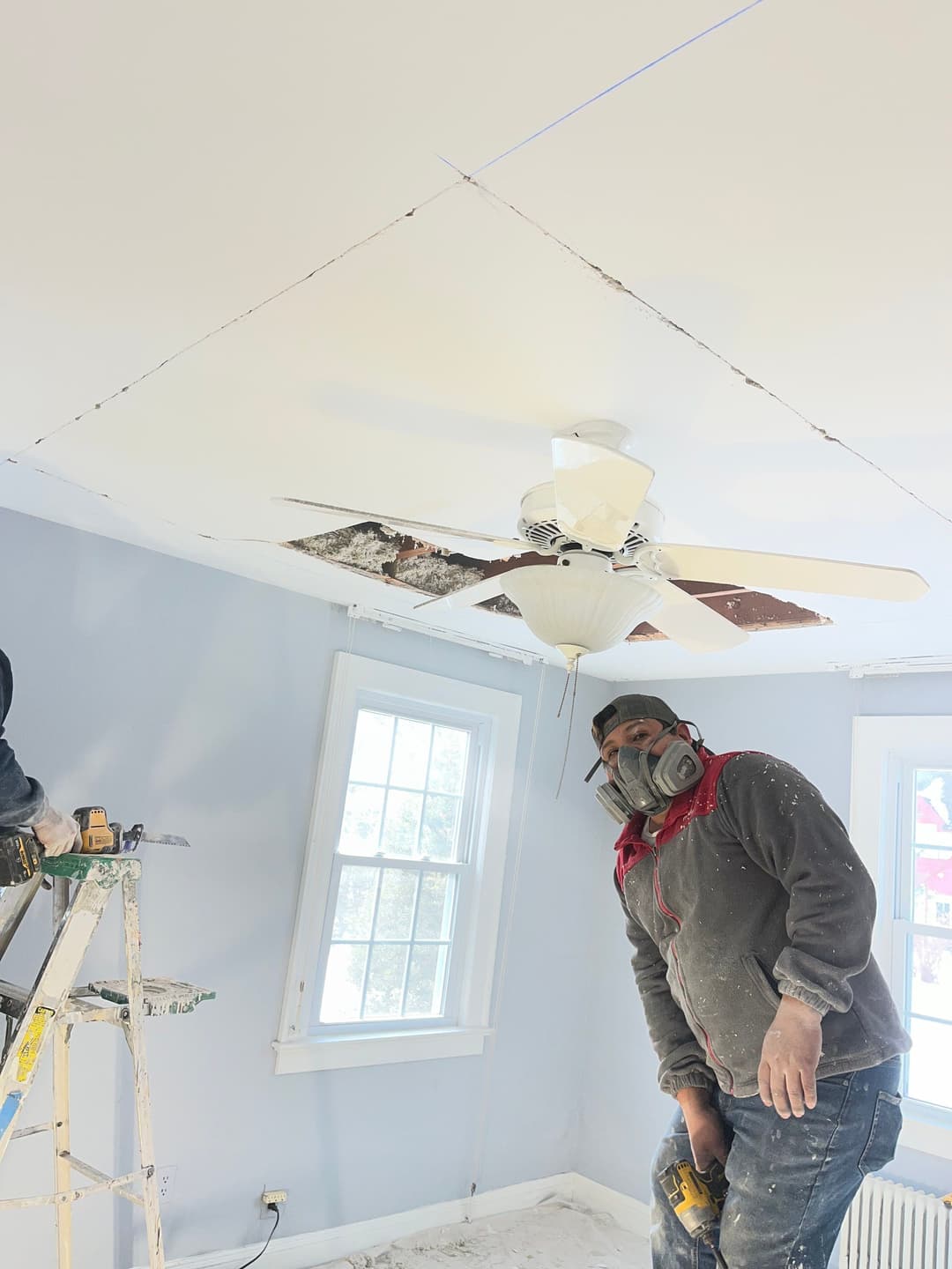 Construction worker wearing a mask, repairing a ceiling with a fan in a light blue room.