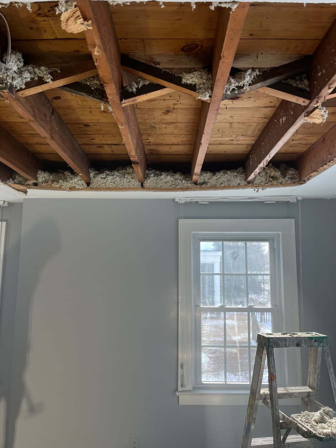 Exposed ceiling beams with insulation in a room under renovation, featuring a ladder nearby.